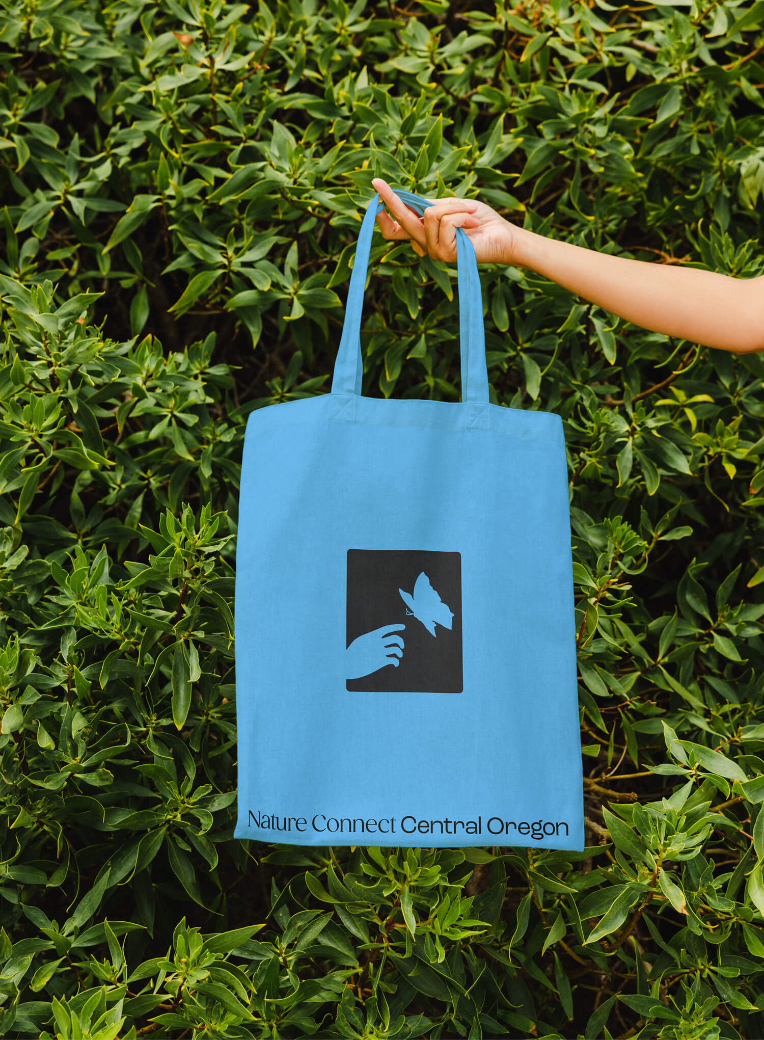 A bright blue tote bag featuring a black square logo with a hand reaching for a butterfly, plus the text ‘Nature Connect Central Oregon,’ held up in front of lush green foliage.