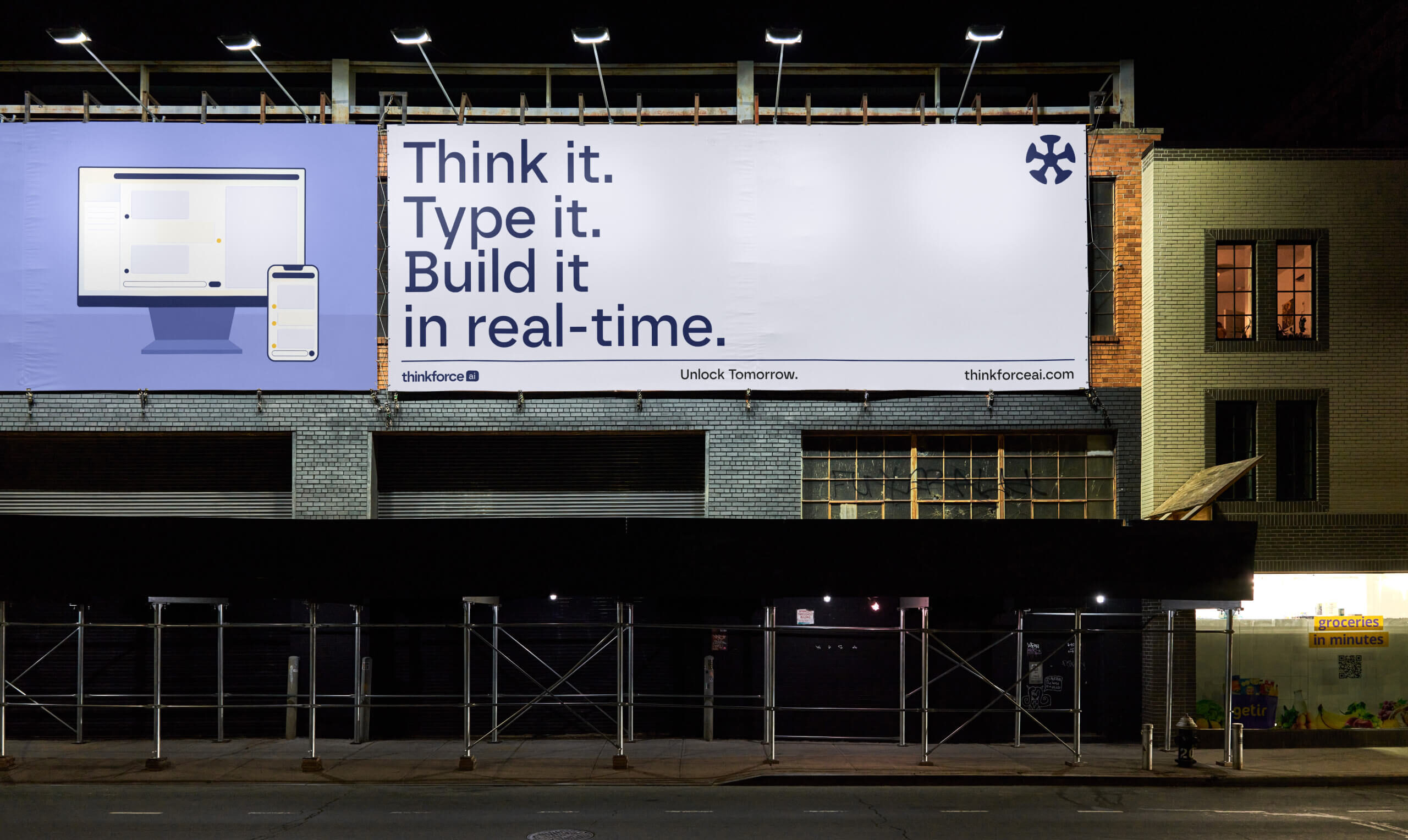 A large outdoor billboard at night promoting the AI platform. The ad reads, “Think it. Type it. Build it in real-time,” with an illustration of a computer and smartphone screen.